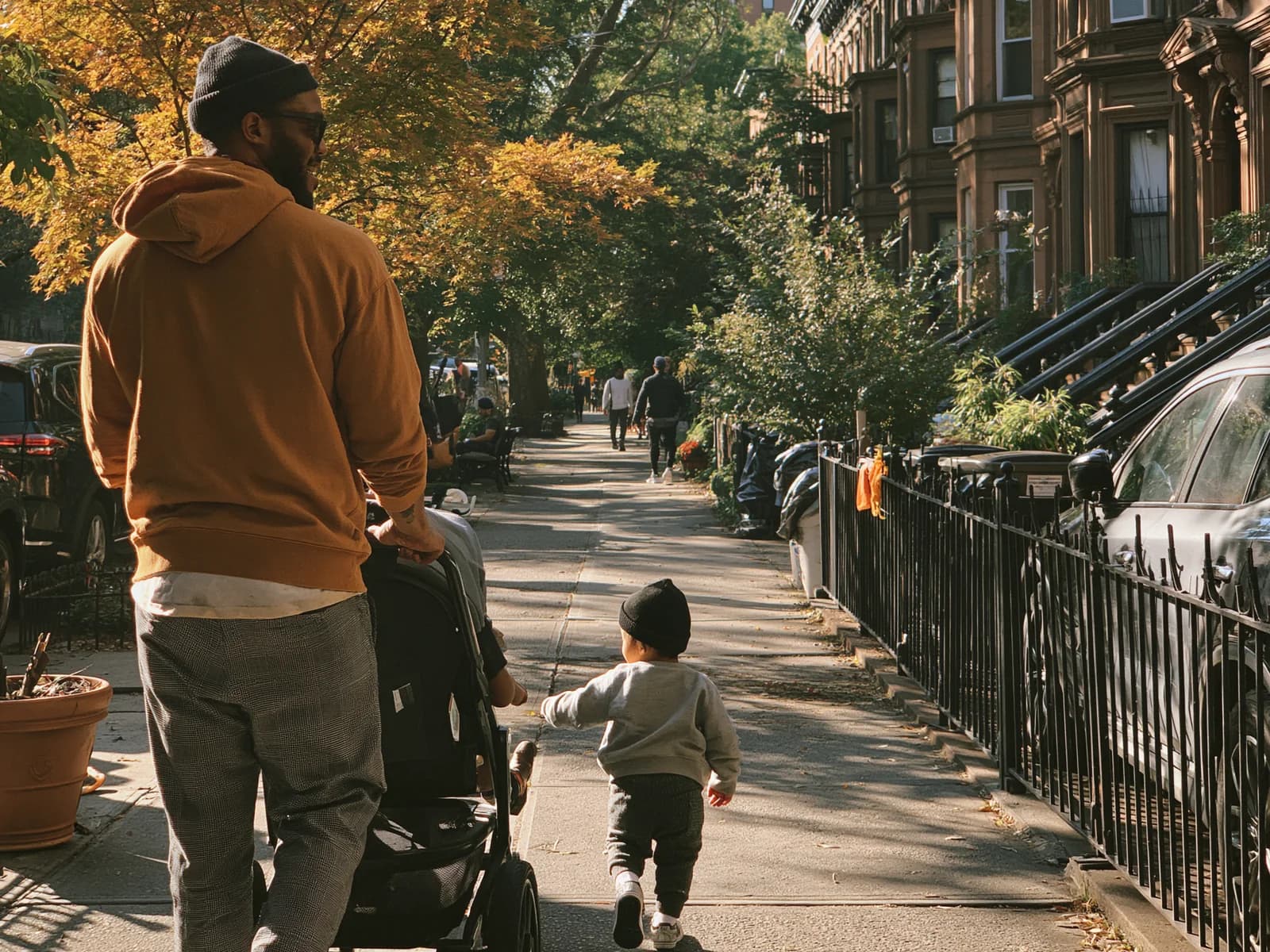 A father pushing a stroller down a Brooklyn brownstone block on an autumn afternoon, a toddler running ahead